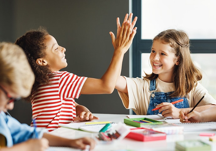 Zwei Mädchen lächeln und geben sich gegenseitig ein High Five, während sie an einem Tisch mit Schulsachen, Heften und Buntstiften sitzen. Ein Junge im Vordergrund konzentriert sich auf seine Arbeit.