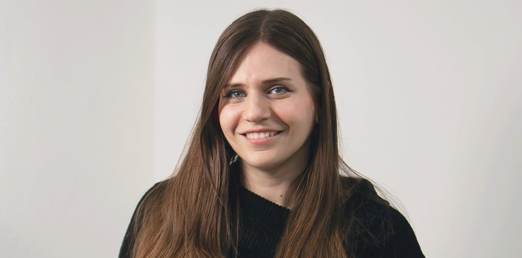 Eine Frau mit langen braunen Haaren, blauen Augen und einem leichten Lächeln, die ein schwarzes Oberteil trägt, steht vor einem schlichten hellen Hintergrund.